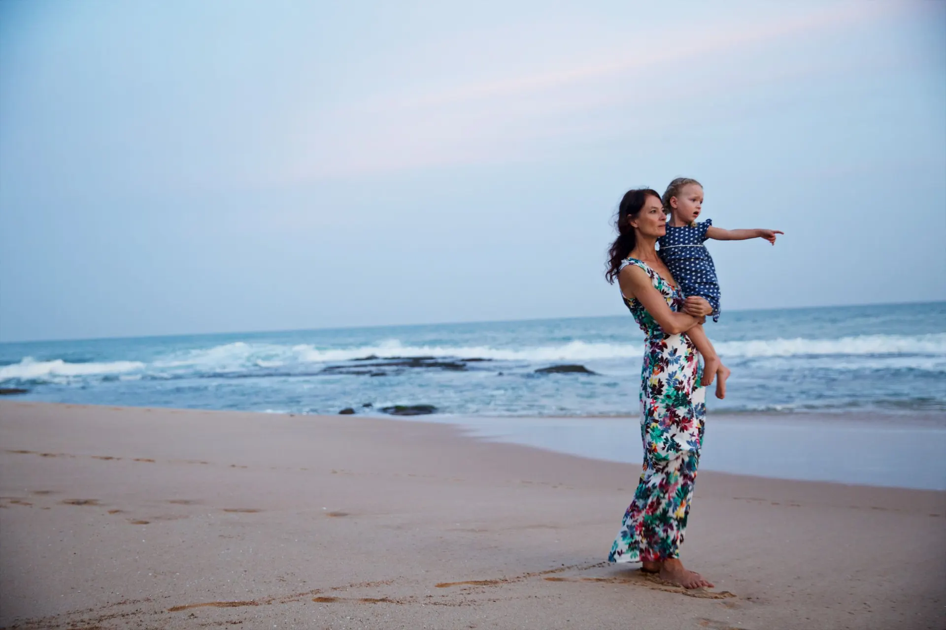 A mother holding her daughter on the beach, pointing out to sea